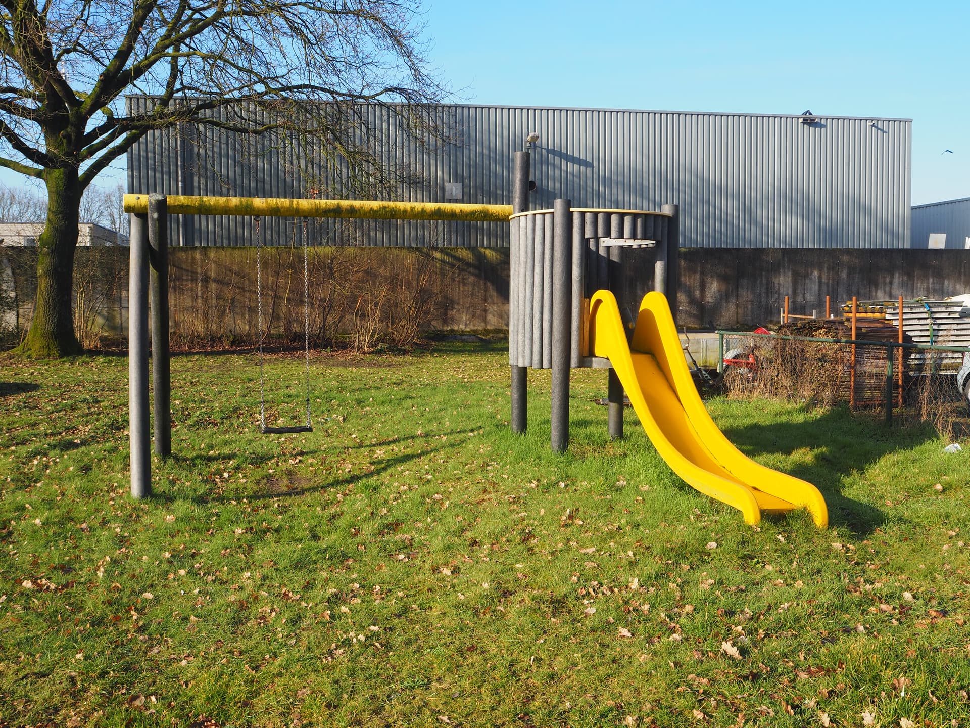 Enlarge image: on the left a swing with a slide to its right, in the background is a commercial building