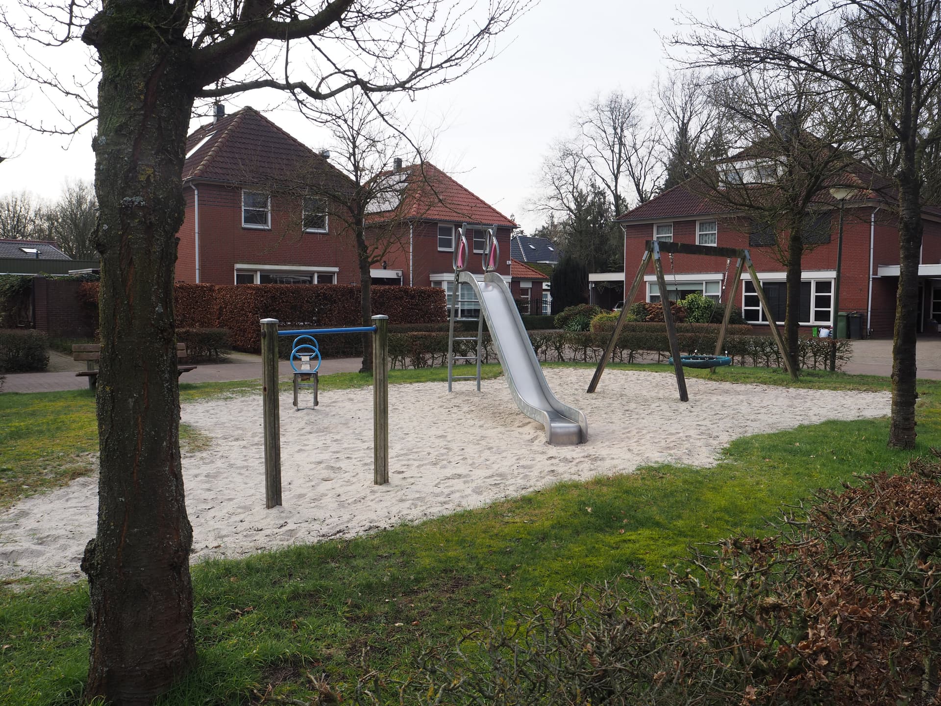 Enlarge image: on the left side a ducking frame and seesaw, to the right of which is a slide and next to it a swing. In the background are houses.